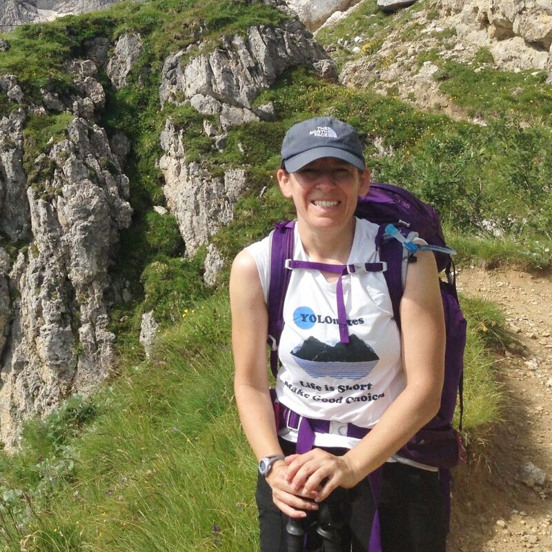 The image shows a woman smiling while hiking on a mountain trail. She is wearing a cap, a white shirt with a graphic, and a backpack. The background features a rocky cliff and green vegetation. She is holding hiking poles.